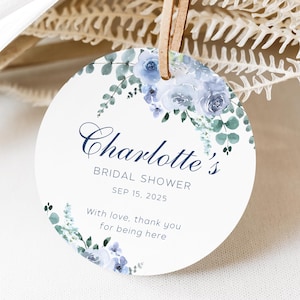 May include: A round, white tag with a blue and green floral design. The tag says "Charlotte's Bridal Shower Sep 15, 2025" and "With love, thank you for being here."
