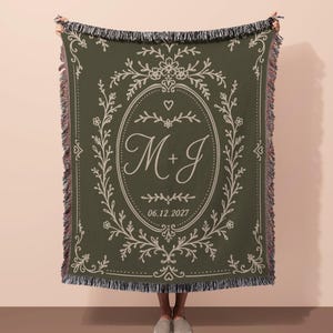 May include: An olive green woven throw blanket featuring a cream-colored floral oval frame with the letters M+J and the date 06.12.2027 printed in the center. The blanket has a decorative fringe along the top and bottom edges with a textured weave.