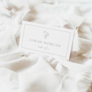 May include: A white rectangular folded place card featuring a thin black border, a small line-art floral illustration, and the printed text Sarah Morgan above the script text Table One, centered on the front of the card with a clean design.