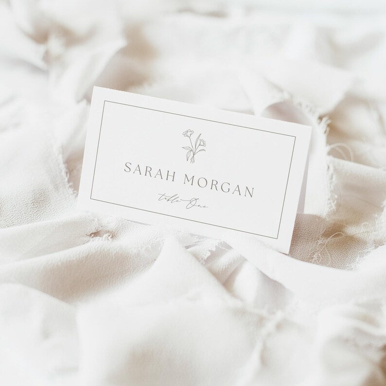 May include: A white rectangular folded place card featuring a thin black border, a small line-art floral illustration, and the printed text Sarah Morgan above the script text Table One, centered on the front of the card with a clean design.