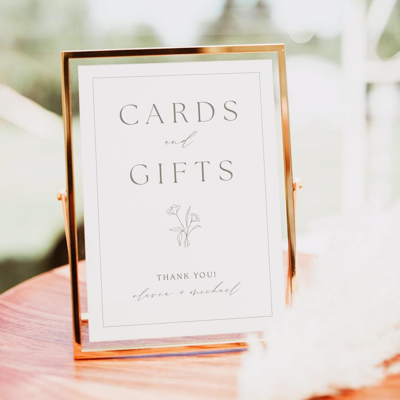 May include: A white sign with a gold frame that says "Cards and Gifts" with a simple floral design and the text "Thank You! Oliver + Michael".