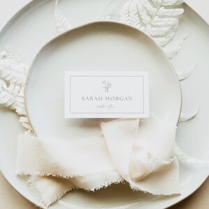 May include: A rectangular white place card with a thin black border, a small line-art floral illustration, and the printed text SARAH MORGAN and table One. The card is centered on a white ceramic plate with a frayed cream-colored fabric ribbon.