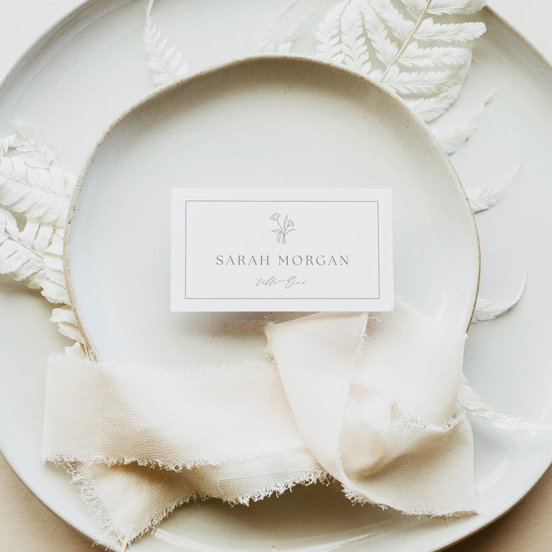 May include: A rectangular white place card with a thin black border, a small line-art floral illustration, and the printed text SARAH MORGAN and table One. The card is centered on a white ceramic plate with a frayed cream-colored fabric ribbon.