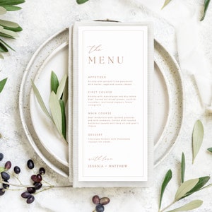 K&ouml;nnte beinhalten: Eine Hochzeitskarte mit einem schlichten, eleganten Design. Das Men&uuml; hat einen wei&szlig;en Hintergrund mit goldener Schrift und einem braunen Rand. Das Men&uuml; umfasst Vorspeisen, ersten Gang, Hauptgericht und Dessert. Der Text lautet: "the MENU", "APPETIZER", "Ricotta und Spinat gef&uuml;llte Panzerotti mit Butter, Salbei und Casera-K&auml;se", "FIRST COURSE", "Risotto mit Mascarpone und trocken gesalzenem Rindfleisch. Serviert mit gemischtem Gr&uuml;n, Zucchini, Gurke, marinierten Paprikaschoten und Zitronen-Vinaigrette", "MAIN COURSE", "Rinderfilet mit sautierten Kartoffeln und wildem Rosmarin. Serviert mit ger&ouml;stetem Butternussk&uuml;rbis mit Kr&auml;uter&ouml;l und Ziegenk&auml;se", "DESSERT", "Schokoladenfondant mit hausgemachtem Kokosnuss-Eis.", "with love", "JESSICA MATTHEW".