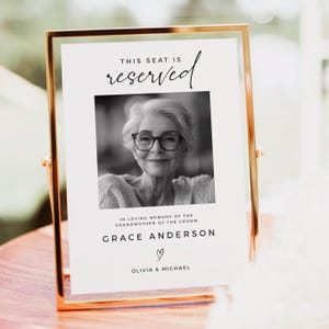 Pode incluir: Uma fotografia preto e branco de uma mulher de óculos está em uma placa branca com texto preto que diz "Este lugar é reservado" e "Em memória carinhosa da avó do noivo Grace Anderson" com um símbolo de coração e os nomes Olivia & Michael.