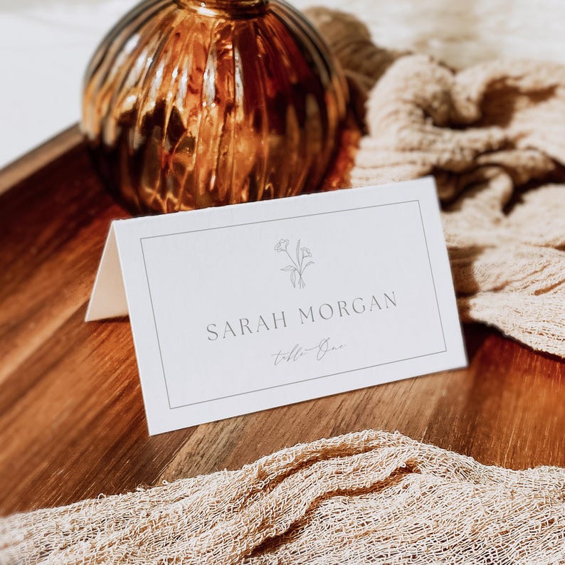 May include: A white folded place card with a thin rectangular border, a small line-art floral illustration, and the printed text "SARAH MORGAN" followed by "Table One" in a script font, positioned on a wooden surface with a textured fabric.