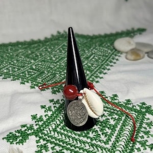 May include: A red cord bracelet featuring a round, polished red bead, a white cowrie shell, and a circular silver-toned metal charm with intricate embossed detailing. The bracelet is displayed on a black, cone-shaped jewellery stand.