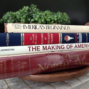 May include: A stack of four hardcover books with visible titles including America's Beginnings, So Proudly We Hail, The Making of America, and The Discovery of North America. The books feature cream, blue, white, and deep red spines with gold lettering.