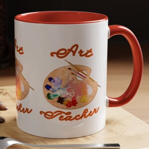 Puede incluir: Taza de café de cerámica blanca con borde y asa naranja. La taza presenta una paleta de pintura colorida con pinceles y el texto "Art Teacher" en naranja.