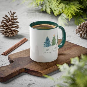 Op de afbeelding: Witte keramische mok met een groene rand en handvat. De mok heeft een aquarel-illustratie van drie kerstbomen met de tekst "Have Yourself a Merry Christmas".