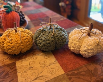 May include: Three crocheted pumpkins in shades of yellow, green, and cream sit on a patterned tablecloth. Each pumpkin has a brown stem and is tied with twine. The pumpkins are a fall-themed decoration.