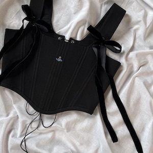 May include: A black corset top featuring vertical structural seams, wide shoulder straps adorned with long black velvet ribbon ties, and a small embroidered orb logo on the chest. The garment includes a front hook-and-eye closure and thin laces.