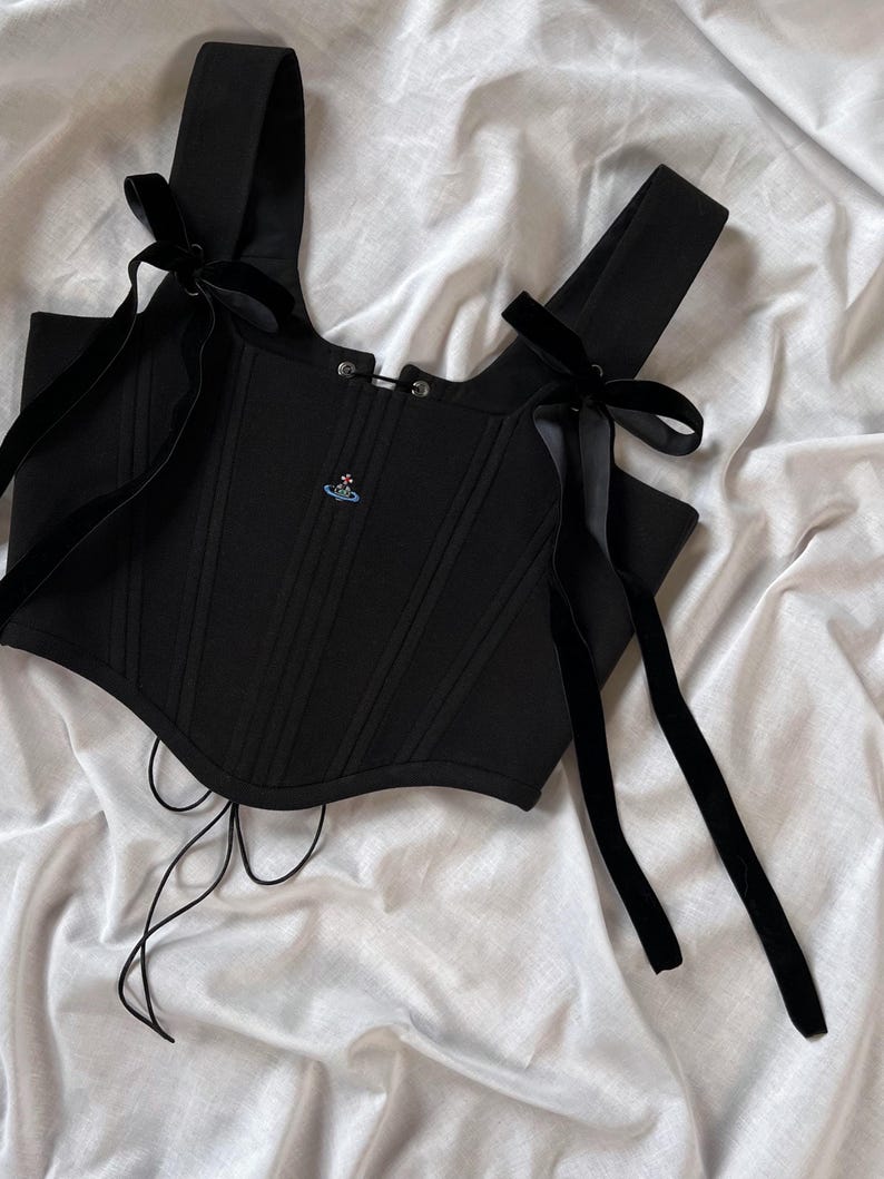 May include: A black corset top featuring vertical structural seams, wide shoulder straps adorned with long black velvet ribbon ties, and a small embroidered orb logo on the chest. The garment includes a front hook-and-eye closure and thin laces.