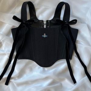 May include: A black corset top featuring a ribbed texture, a lace-up front detail, and wide shoulder straps adorned with long black velvet ribbons tied into bows. A small, embroidered blue and silver orb logo is centered on the chest.