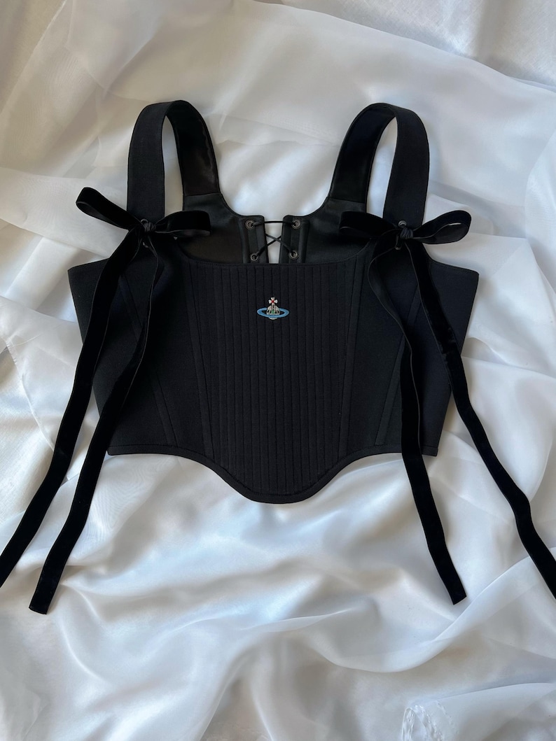 May include: A black corset top featuring a ribbed texture, a lace-up front detail, and wide shoulder straps adorned with long black velvet ribbons tied into bows. A small, embroidered blue and silver orb logo is centered on the chest.