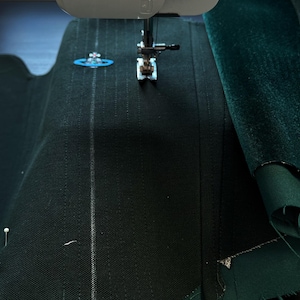 May include: A sewing machine needle positioned over dark green fabric with a textured weave. The fabric features vertical stitched lines and a small, embroidered light blue orb logo with a pink cross detail on the upper left side.