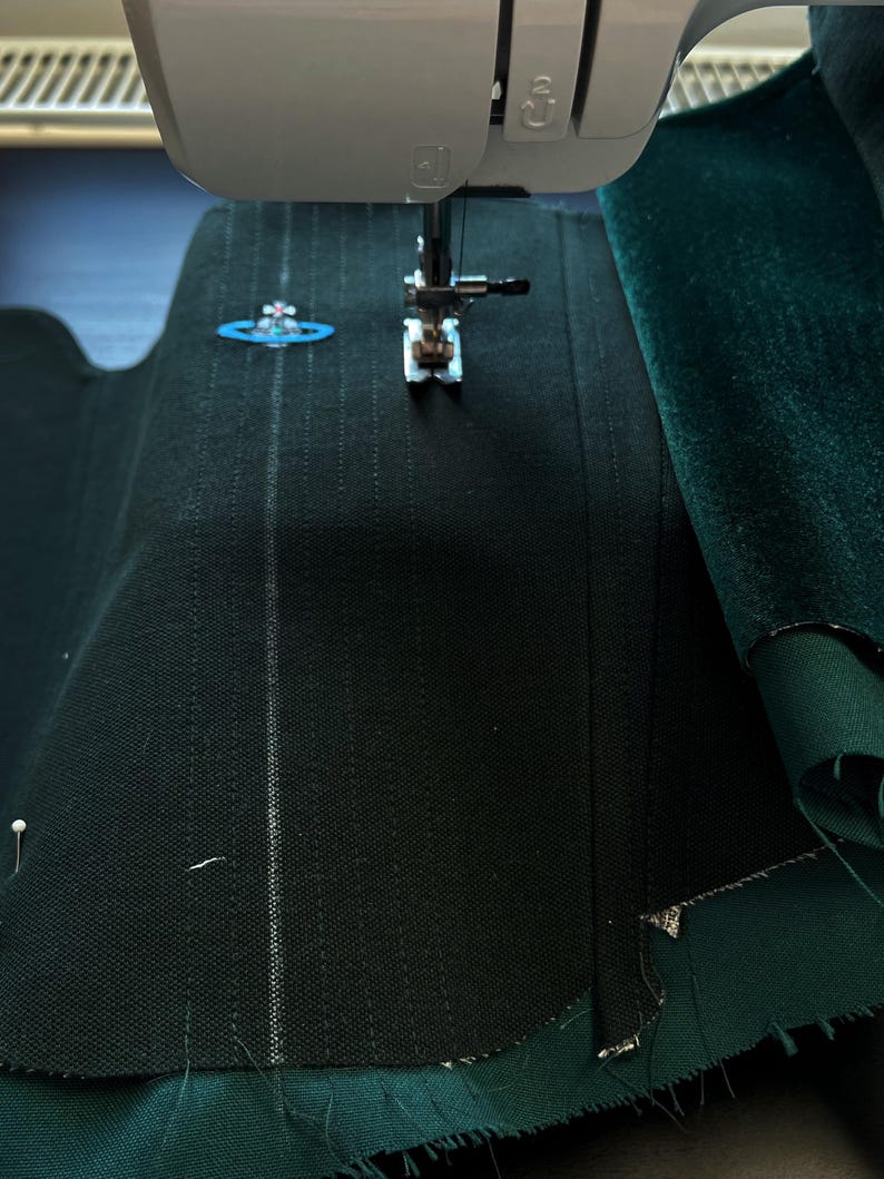 May include: A sewing machine needle positioned over dark green fabric with a textured weave. The fabric features vertical stitched lines and a small, embroidered light blue orb logo with a pink cross detail on the upper left side.