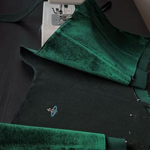 May include: A dark green velvet fabric piece being sewn on a sewing machine. The fabric features a small embroidered logo of a planet with a ring and a cross on top. The material has a soft, reflective texture and is pinned in several places.