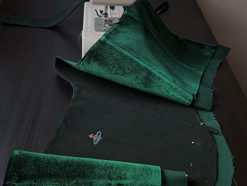 May include: A dark green velvet fabric piece being sewn on a sewing machine. The fabric features a small embroidered logo of a planet with a ring and a cross on top. The material has a soft, reflective texture and is pinned in several places.