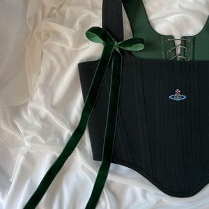 May include: A black corset top featuring a dark green velvet ribbon bow on the shoulder strap. The garment has a square neckline, a lace-up front closure with metal eyelets, and a small embroidered orb logo in blue, red, and silver on the front.