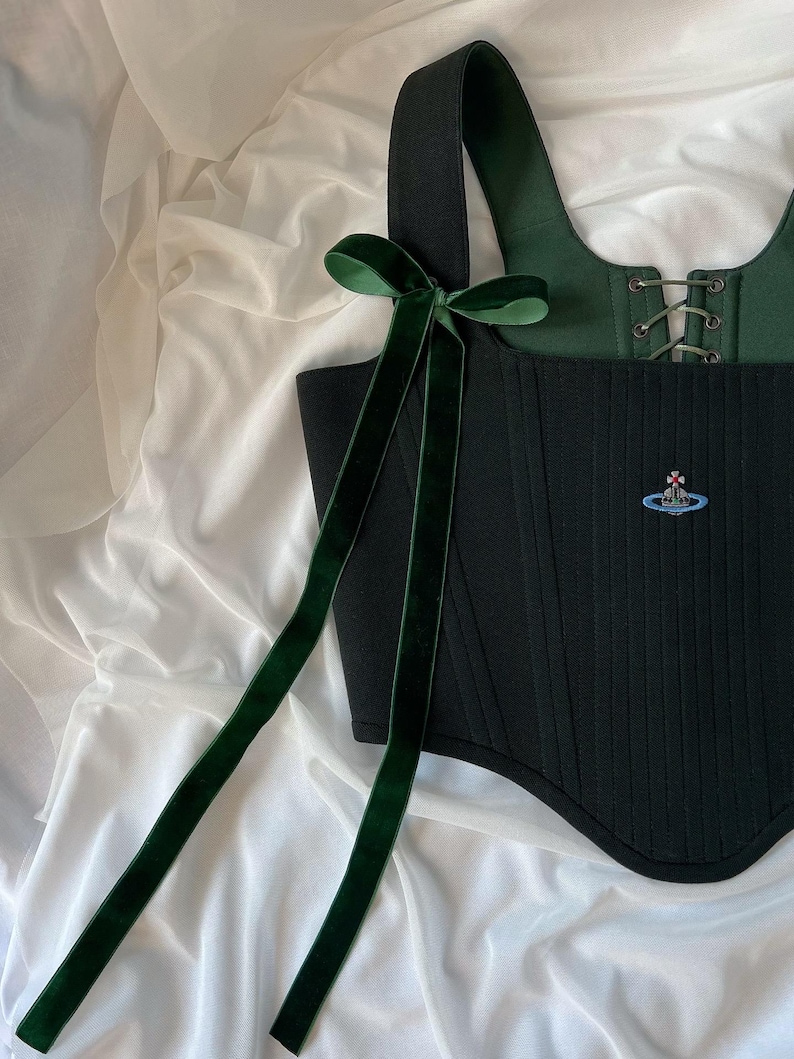 May include: A black corset top featuring a dark green velvet ribbon bow on the shoulder strap. The garment has a square neckline, a lace-up front closure with metal eyelets, and a small embroidered orb logo in blue, red, and silver on the front.