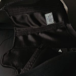 May include: A black satin corset under construction, featuring visible stitched seams and a rectangular white fabric label sewn onto the interior. The label displays the text Vivienne Westwood MAN with a small orb logo above the brand name.