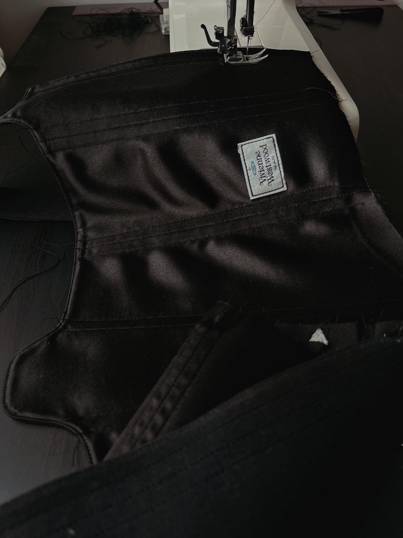 May include: A black satin corset under construction, featuring visible stitched seams and a rectangular white fabric label sewn onto the interior. The label displays the text Vivienne Westwood MAN with a small orb logo above the brand name.