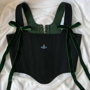 May include: A black corset top featuring wide shoulder straps adorned with long dark green velvet ribbons tied into bows. The bodice includes a central lace-up closure with metal eyelets, a dark green interior lining, and a small embroidered orb logo.