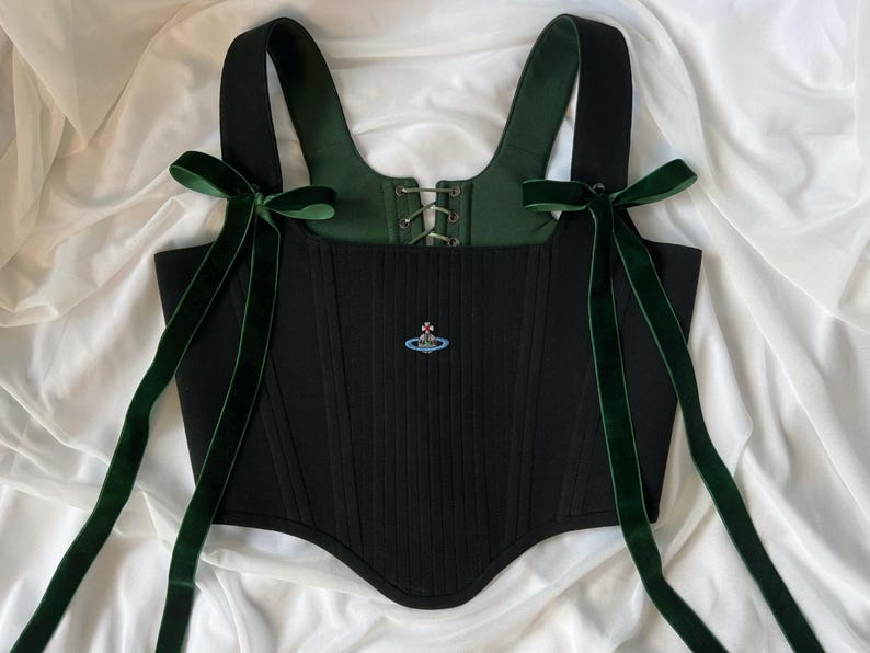 May include: A black corset top featuring wide shoulder straps adorned with long dark green velvet ribbons tied into bows. The bodice includes a central lace-up closure with metal eyelets, a dark green interior lining, and a small embroidered orb logo.
