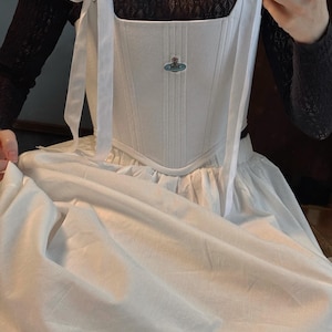 May include: A white corset-style bodice featuring vertical stitching, ribbon shoulder ties, and a small embroidered orb logo on the chest. The bodice is worn over a black long-sleeved lace top and paired with a full, plain white skirt.
