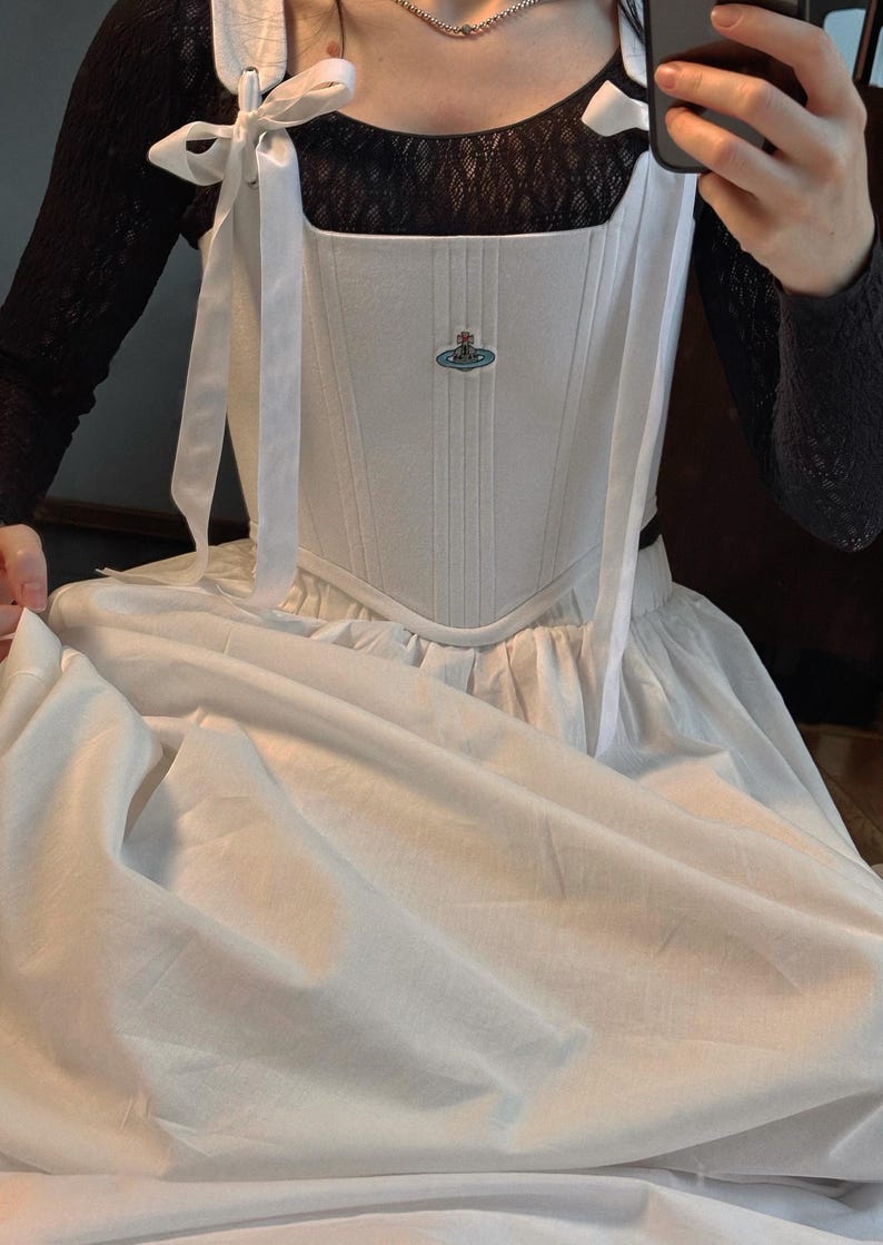 May include: A white corset-style bodice featuring vertical stitching, ribbon shoulder ties, and a small embroidered orb logo on the chest. The bodice is worn over a black long-sleeved lace top and paired with a full, plain white skirt.