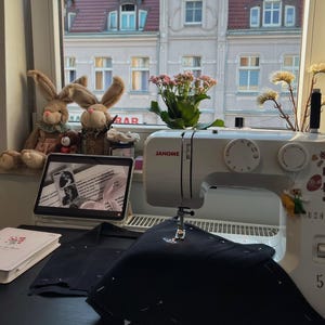 May include: A white Janome sewing machine sits on a black surface with black fabric pinned under the needle. A tablet displaying text, two plush rabbit toys, a small potted plant, and a white notebook are arranged nearby on the windowsill.