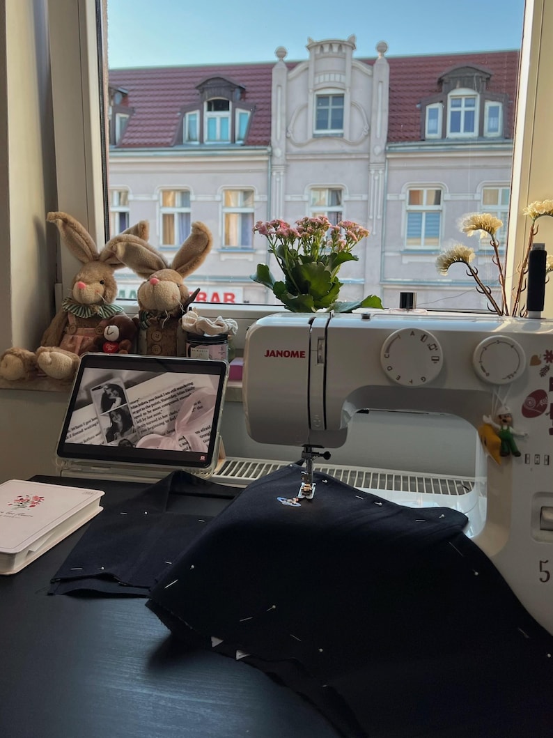 May include: A white Janome sewing machine sits on a black surface with black fabric pinned under the needle. A tablet displaying text, two plush rabbit toys, a small potted plant, and a white notebook are arranged nearby on the windowsill.