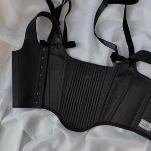 May include: A black corset top featuring vertical pleats, a side panel with silver-toned eyelets, and adjustable shoulder straps. A white rectangular label with the text Vivienne Westwood MAN is stitched onto the lower right corner of the garment.