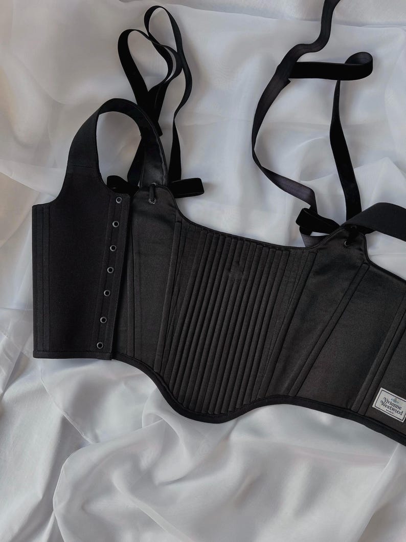 May include: A black corset top featuring vertical pleats, a side panel with silver-toned eyelets, and adjustable shoulder straps. A white rectangular label with the text Vivienne Westwood MAN is stitched onto the lower right corner of the garment.