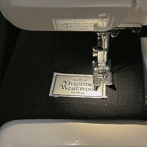 May include: A black fabric garment featuring a white rectangular label with the text Vivienne Westwood MAN and an orb logo, positioned under the metal presser foot and needle of a white sewing machine, with a partial label visible above.