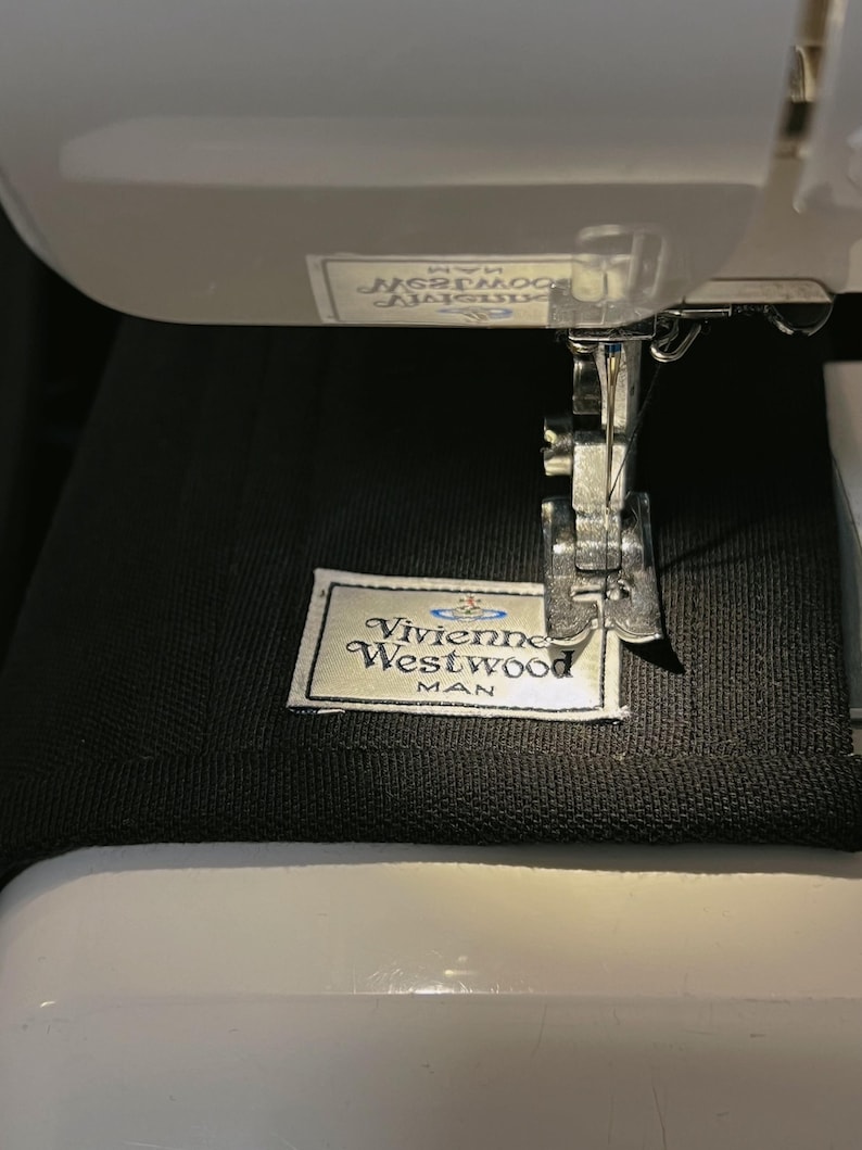 May include: A black fabric garment featuring a white rectangular label with the text Vivienne Westwood MAN and an orb logo, positioned under the metal presser foot and needle of a white sewing machine, with a partial label visible above.