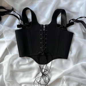 May include: A black corset top featuring a front lace-up closure with black eyelets and thin black cord ties. The garment includes wide shoulder straps with adjustable ribbon ties and a structured, paneled bodice with a curved hemline at the waist.