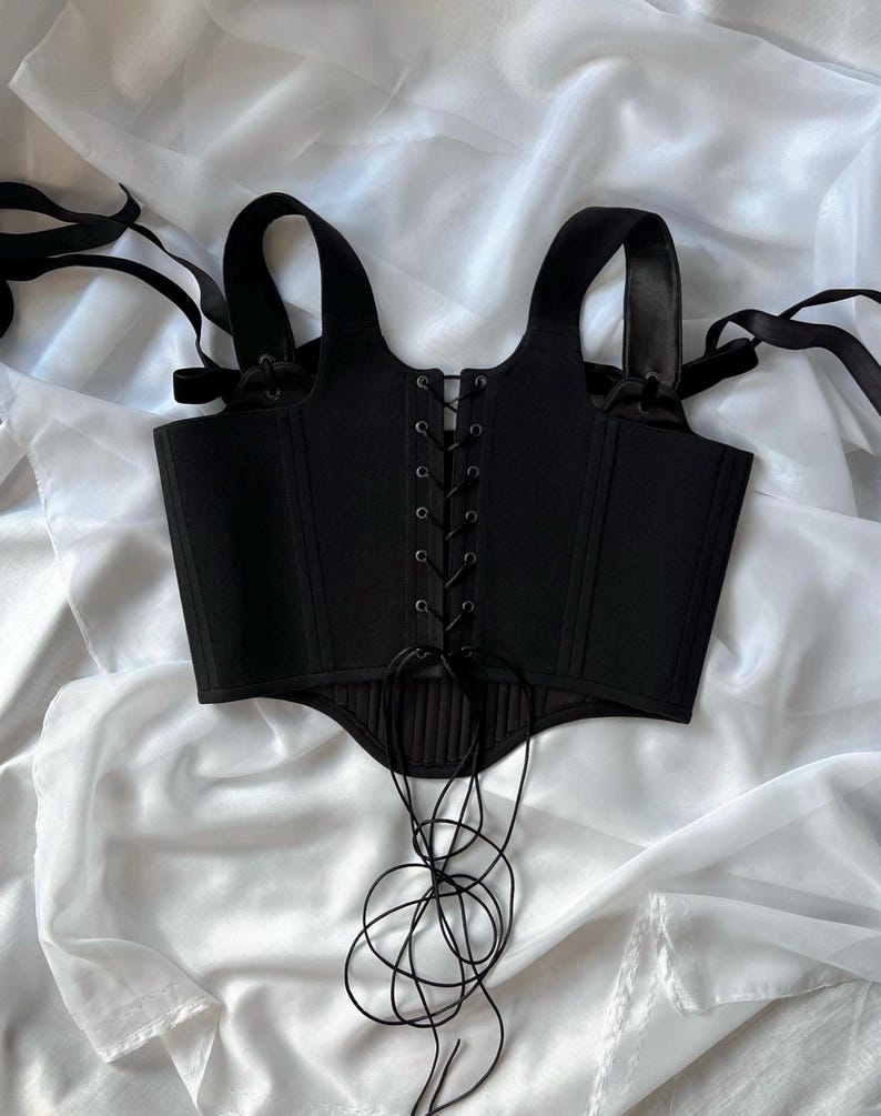 May include: A black corset top featuring a front lace-up closure with black eyelets and thin black cord ties. The garment includes wide shoulder straps with adjustable ribbon ties and a structured, paneled bodice with a curved hemline at the waist.