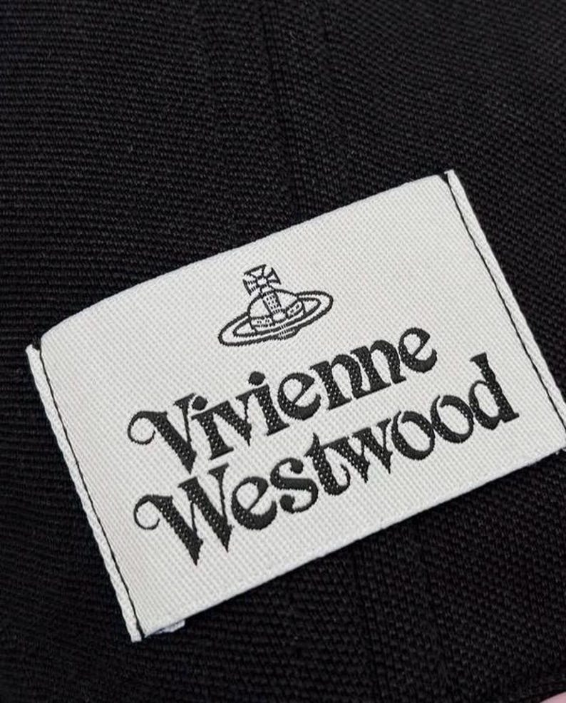 May include: A rectangular white fabric label stitched onto a black textured fabric background. The label features the black embroidered text Vivienne Westwood in a stylized serif font, positioned below a black embroidered orb and cross logo.