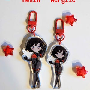 K&ouml;nnte beinhalten: Zwei Versionen eines durchsichtigen Acryl-Schl&uuml;sselanh&auml;ngers mit einer Comicfigur. Der Schl&uuml;sselanh&auml;nger ist 7,6 cm hoch in Resin und 6,4 cm hoch in Acryl. Die Figur ist eine Frau mit schwarzen Haaren und einem rot-schwarzen Outfit. Der Schl&uuml;sselanh&auml;nger hat einen roten Metallverschluss.