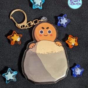Könnte beinhalten: Ein klarer Acryl-Schlüsselanhänger mit einer Cartoonfigur mit orangefarbenem Kopf und grauem Körper. Der Schlüsselanhänger hat einen goldfarbenen Verschluss. Mehrere sternförmige Charms in Blau und Orange umgeben den Schlüsselanhänger. Der Text "Digi's Charms" ist ebenfalls sichtbar.