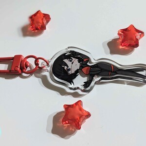 K&ouml;nnte beinhalten: Ein durchsichtiger Acryl-Schl&uuml;sselanh&auml;nger mit einer Comicfigur mit schwarzen Haaren, einem rot-schwarzen Outfit und einem roten Augenpflaster. Der Schl&uuml;sselanh&auml;nger hat einen silbernen Metallring und einen roten Metallverschluss.