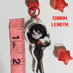 K&ouml;nnte beinhalten: Ein transparenter Acryl-Anh&auml;nger mit einem schwarzen und roten Anime-Charakter in einem rot-schwarzen Outfit. Der Anh&auml;nger ist an einem roten Metall-Karabinerhaken befestigt. Der Anh&auml;nger ist etwa 5 cm lang. Der Anh&auml;nger wird auf einem rosa Lineal gezeigt.