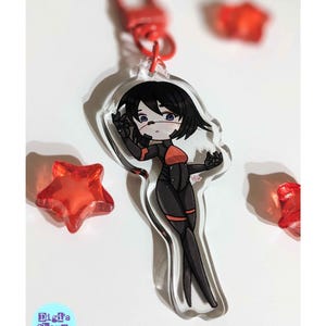K&ouml;nnte beinhalten: Ein durchsichtiger Acryl-Schl&uuml;sselanh&auml;nger mit einer Comicfigur mit schwarzen Haaren, einem rot-schwarzen Outfit und einem roten Stern auf der Brust. Der Schl&uuml;sselanh&auml;nger ist an einem roten Metallverschluss befestigt.