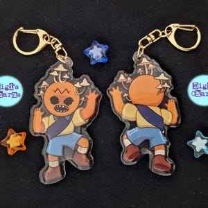 Könnte beinhalten: Zwei Schlüsselanhänger aus Acryl mit einem Cartoon-Charakter mit einem kürbisförmigen Kopf und Pilzdetails. Die Figur trägt ein gelbes Hemd, blaue Shorts und eine blaue Schärpe. Goldfarbene Verschlüsse und sternförmige Charms sind ebenfalls sichtbar.