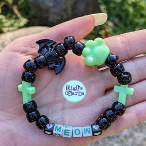 Può includere: Braccialetto di perline nere e verdi con ciondoli a forma di pipistrello, zampa, croce e cuore. Il braccialetto scrive "MEOW" in lettere bianche.