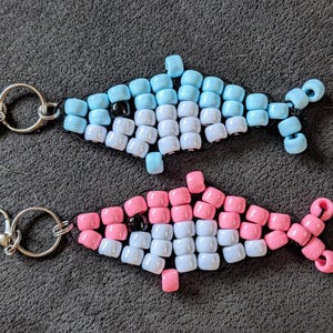 Könnte beinhalten: Zwei Schlüsselanhänger in Fischform aus Perlen. Einer ist hellblau und weiß, der andere rosa und weiß. Beide haben schwarze Augen und silberne Schlüsselringe. Die Fische bestehen aus kleinen, quadratischen Perlen.