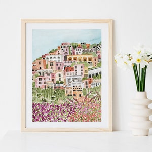 May include: A watercolor painting of a colorful Italian village with a hillside of purple flowers in the foreground. The village is painted in shades of pink, yellow, orange, and green. The painting is framed in a light wood frame.