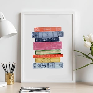 May include: A framed art print featuring a vertical stack of colorful books in shades of orange, blue, pink, green, red, and yellow. The books have decorative spines and text. The print is displayed on a desk with a white lamp, a notebook, and white tulips.